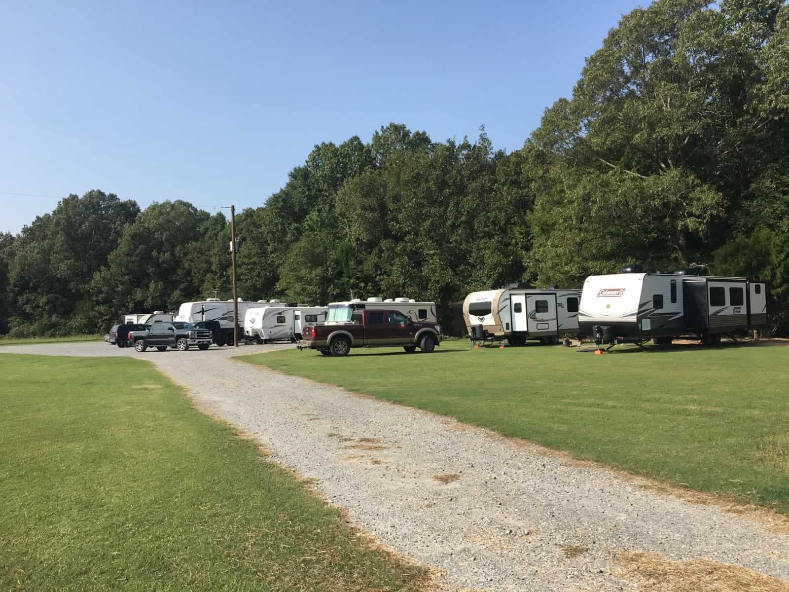 A short gravel road leads to an open lot, where several RVs, campers, and trucks are parked near shady trees.